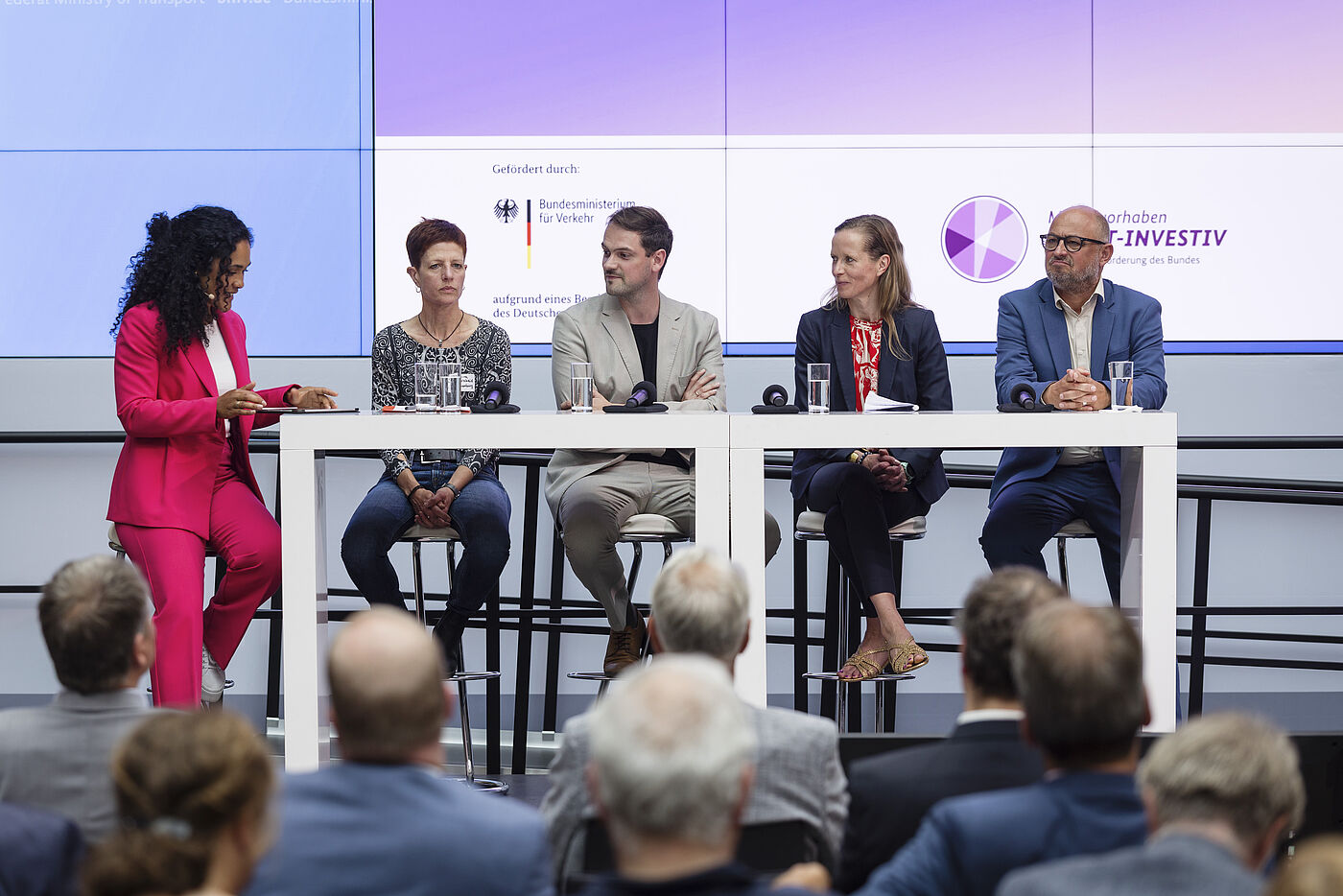 Podiumsdiskussion mit fünf Personen an weißen Tischen vor lila Hintergrund, Moderatorin in pinkem Anzug links, vier Diskussionsteilnehmende rechts, darunter Mann in beigem Anzug und Mann in blauem Anzug ganz rechts, Publikum sitzt davor